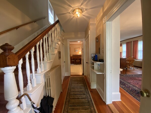 A hallway with a wooden staircase, a radiator, and a rug leading to a room with a desk and chair.
