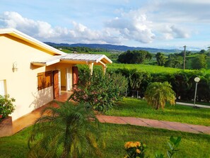 Un bâtiment jaune doté d’un porche abrité, entouré d’une végétation luxuriante et offrant une vue sur des collines lointaines.