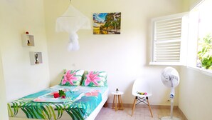 Een slaapkamer met een bed, een klein tafeltje, een stoel, een ventilator en een raam met jaloezieën.
