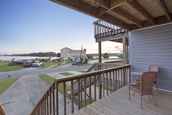 A wooden deck with a railing, a wicker chair, and a view of a street with parked cars and a house in the background. - Adorable Townhome with Water Views (Jacksonville)