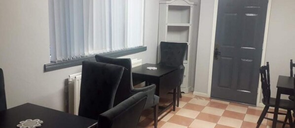 Una habitación con una mesa de comedor negra, dos sillas negras, un radiador blanco, una ventana con persianas blancas, un estante blanco y una puerta negra.