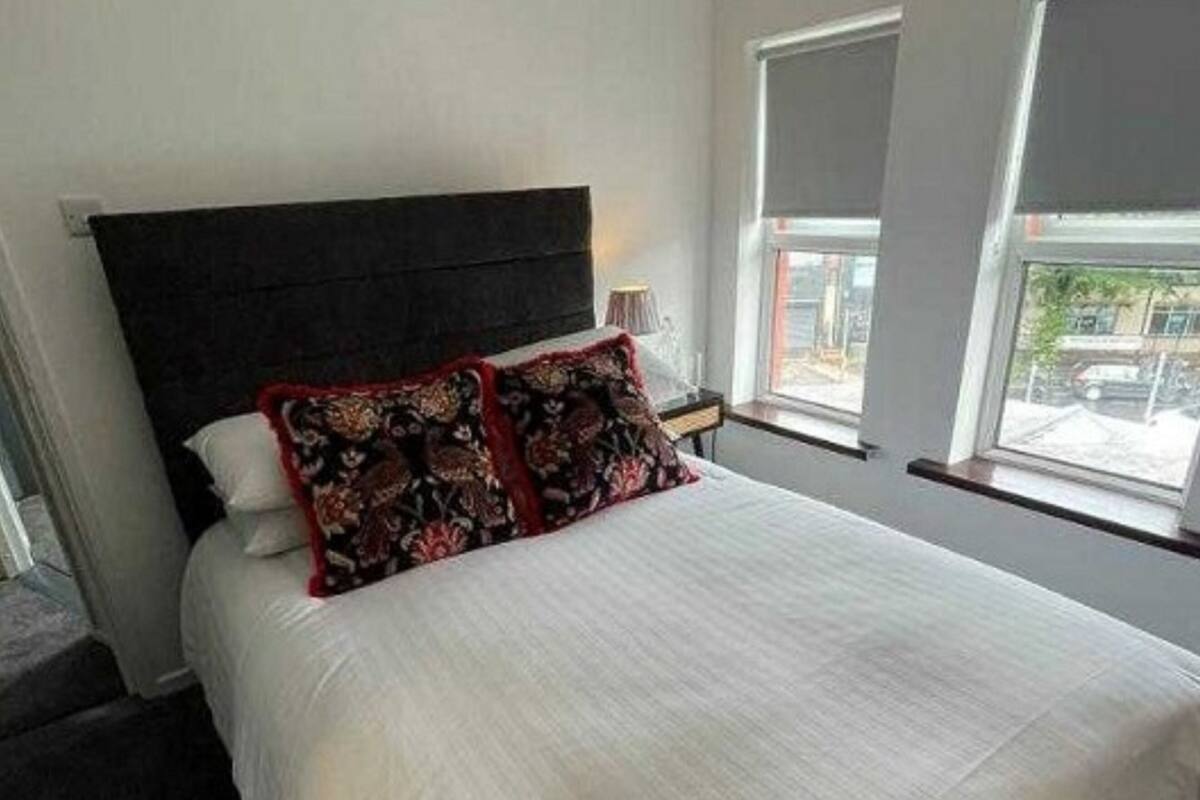 A modern hotel room with a large bed, dark headboard, and two windows providing a view of the outside.