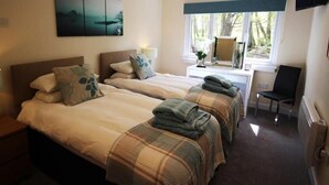 Ett hotellrum med två sängar, ett skrivbord med en stol, ett fönster med utsikt över träd och en tavla på väggen.