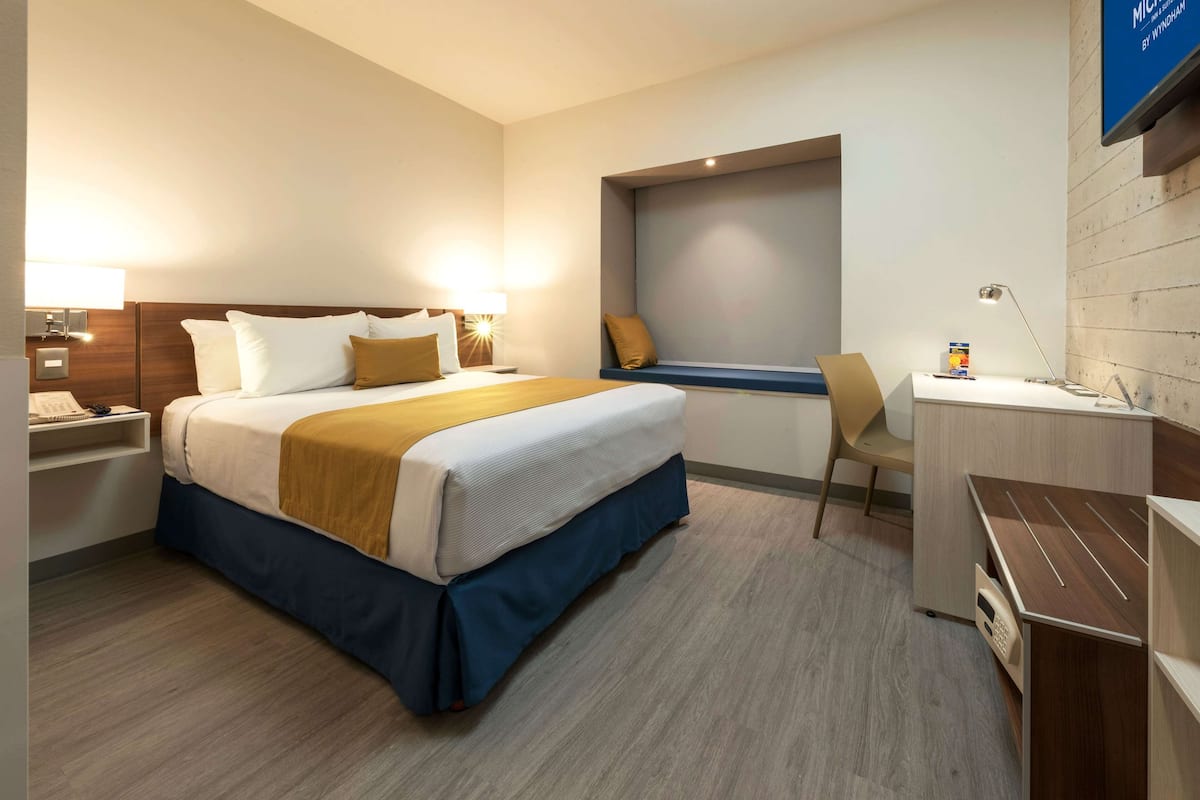 A hotel room with a bed, bedside table, lamp, desk, chair, and a small window.
