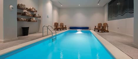 Una piscina interior moderna con agua azul cristalina, sillas de madera y un banco azul.