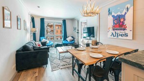 A modern living room with a sofa, dining table, and chairs. There is a TV, a chandelier, and a poster on the wall.
