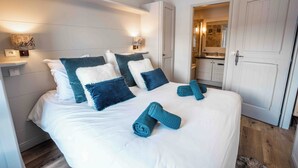 A neatly made bed with white and teal bedding, a pair of rolled towels, and two wall-mounted lamps.