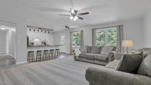 Ein geräumiges Wohnzimmer mit grauem Sofa, Deckenventilator und einer Küche im Hintergrund.