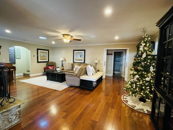Un salón espacioso con pisos de madera, ventilador de techo, un gran árbol de Navidad y chimenea.