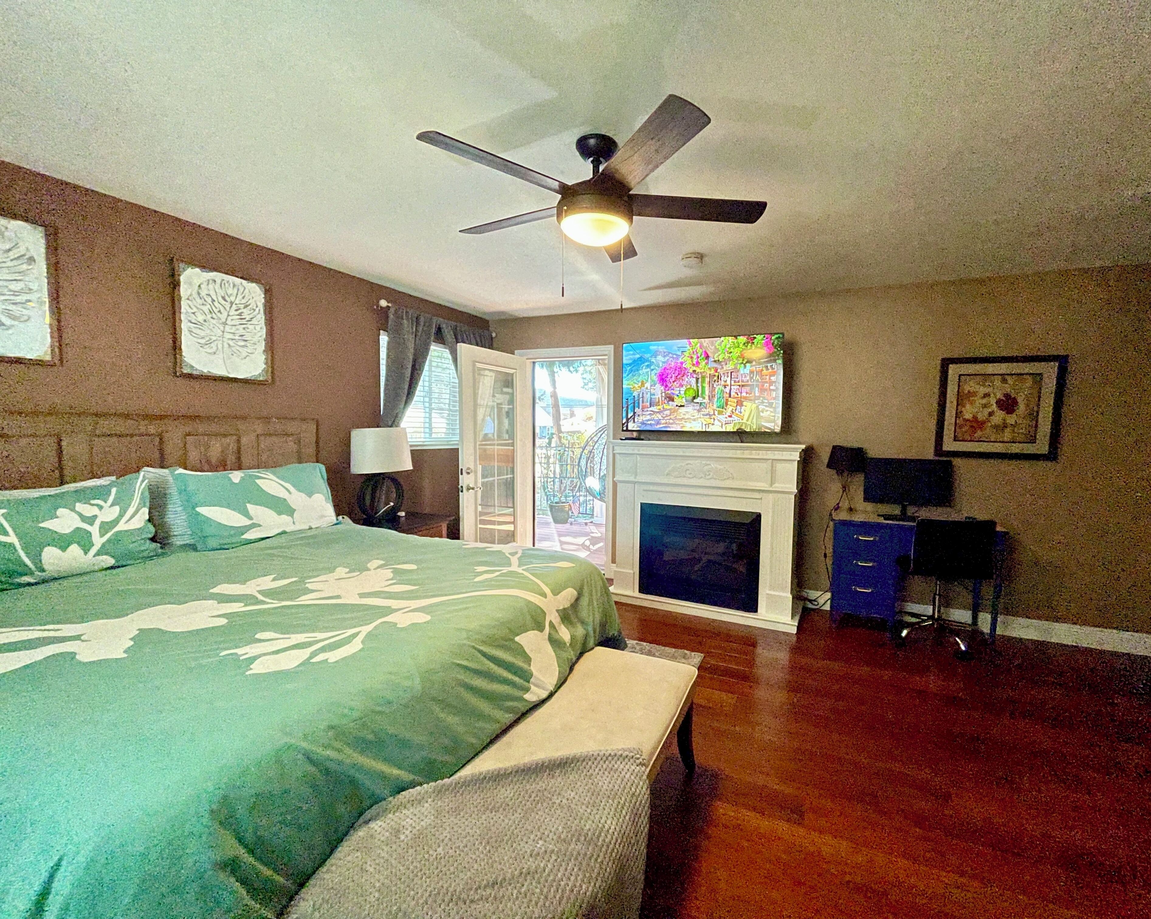 Ein Schlafzimmer mit Bett, Deckenventilator, Kamin, Fernseher und einer Tür, die auf einen Balkon führt.