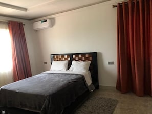 Une chambre d’hôtel avec un lit, une fenêtre avec des rideaux, un appareil de climatisation et un sol recouvert de moquette.
