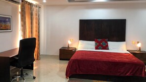 A hotel room with a bed, a desk, a chair, and a nightstand with a lamp.