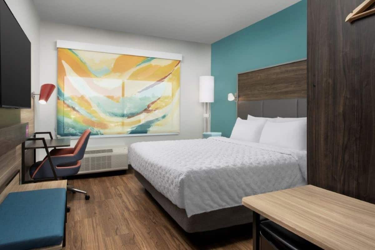 A hotel room with a bed, a desk, a chair, a large window with a painting, and a wall-mounted lamp.