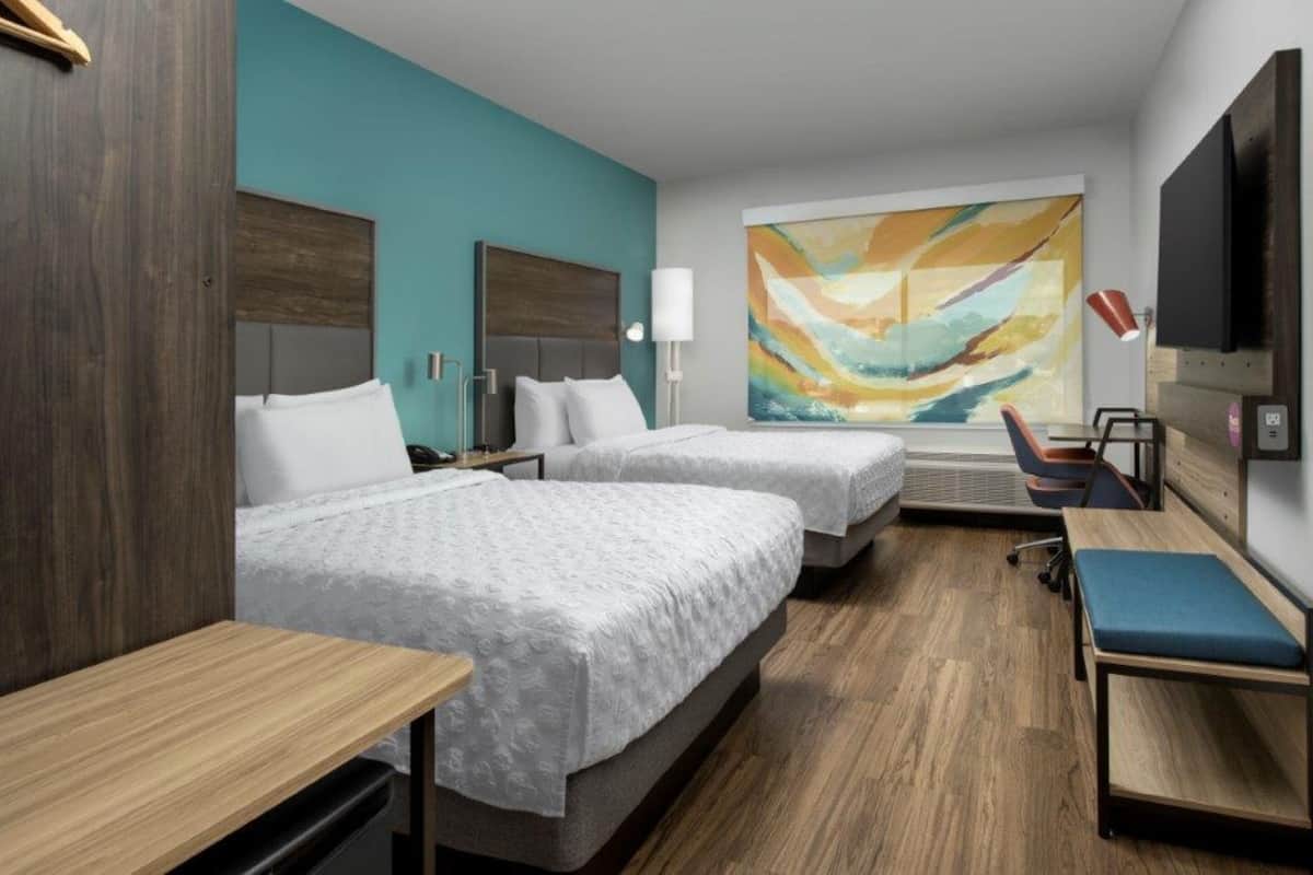 A hotel room with two beds, a desk, a chair, a TV, and a large abstract painting on the wall.