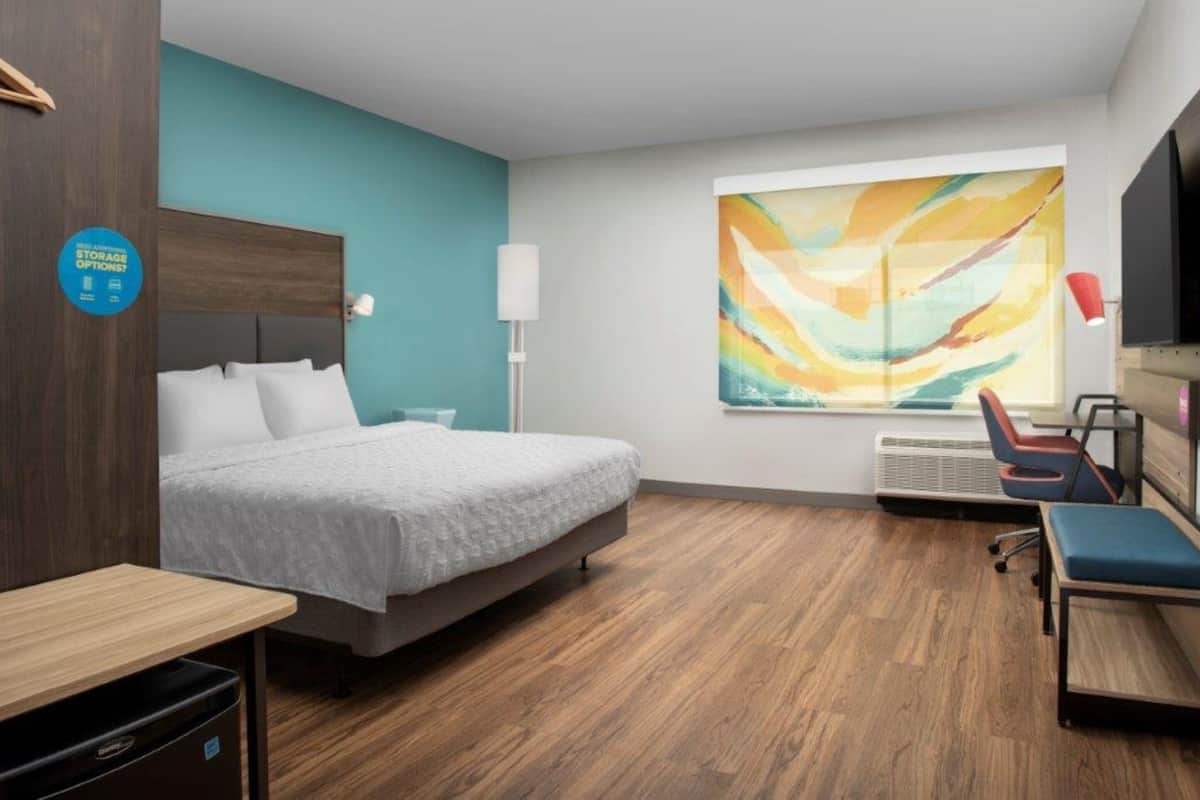A modern hotel room with a bed, a desk, a chair, a TV, and a large abstract painting on the wall.