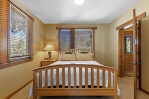 Ein Schlafzimmer mit einem hölzernen Bett, zwei Fenstern mit Jalousien, einer Nachttischlampe und einer Tür zu einem anderen Raum.