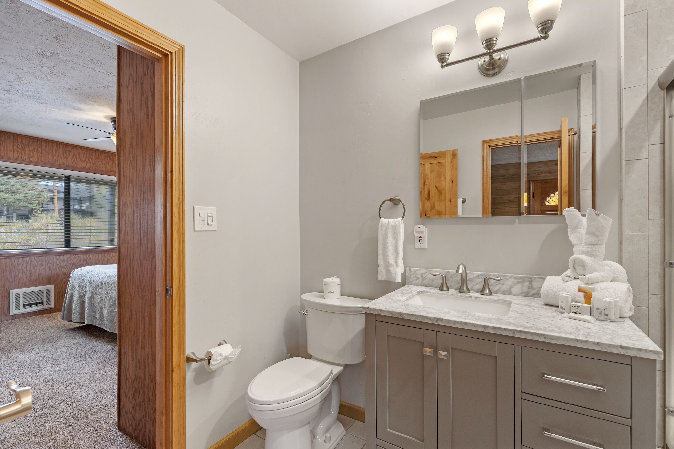 A bathroom with a toilet, sink, and mirror. There is a towel rack above the sink and a window with blinds in the background.