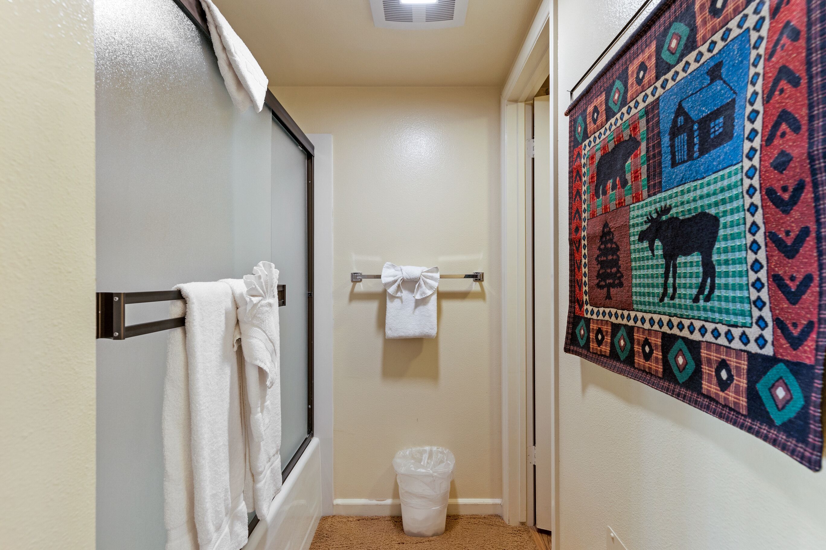 A bathroom with a shower, a towel rack, a trash bin, and a colorful wall decoration.