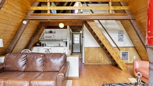 Une pièce de style loft avec un plafond en bois, un canapé en cuir marron, un meuble blanc et un coin cuisine visible à travers une porte ouverte.