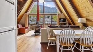 Une pièce mansardée chaleureuse avec un plafond en bois, une grande fenêtre donnant sur une forêt, une cheminée et un coin repas avec des chaises blanches.