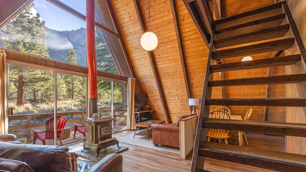 Interior acolhedor de uma cabana, com escada de madeira, lareira e uma grande janela com vista para árvores e montanhas.