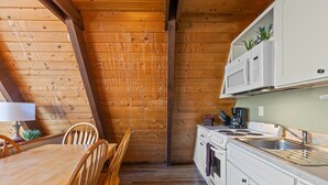 Interior de madeira com uma mesa e cadeiras para refeições, cozinha com armários e eletrodomésticos brancos, e teto de madeira.