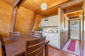 A wooden interior with a dining table and chairs, a kitchen area with white cabinets and appliances, and a door leading outside.