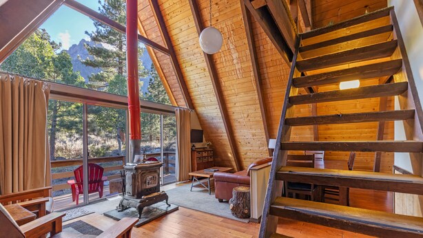 A cozy wooden interior with a loft bedroom, a wooden staircase, and a large window overlooking a forested area.