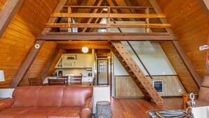 An attic bedroom with a sloped ceiling, wooden beams, a brown leather sofa, a small round table, and a wooden stool.