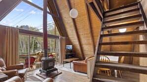 A cozy cabin interior with a wooden staircase, a fireplace, leather furniture, and a large window offering a view of the outdoors. - Four Seasons Cabin 5 (June Lake)