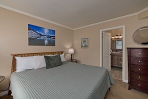 A bedroom with a bed, a nightstand, a lamp, a dresser, a mirror, a painting, and a door leading to a bathroom.