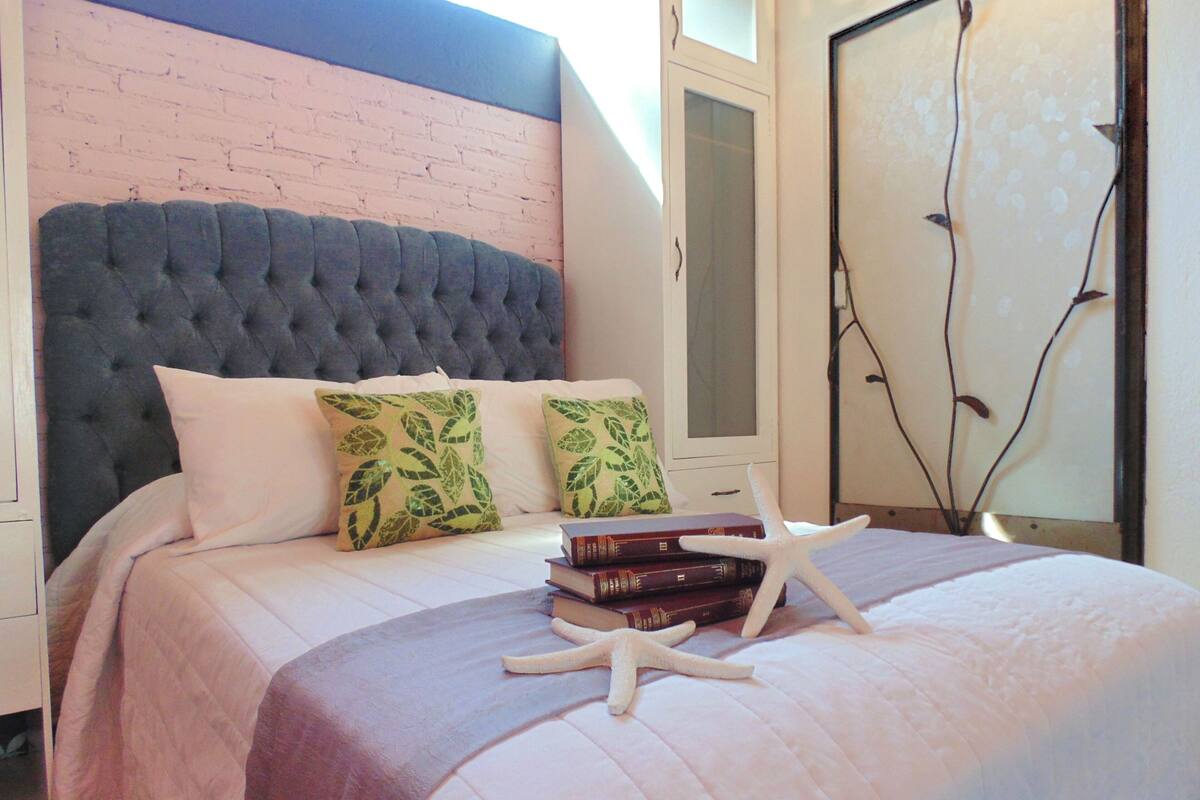 A bedroom with a tufted headboard, a bed with a white and purple bedspread, two pillows with leaf patterns, a stack of books, and a white starfish decoration.