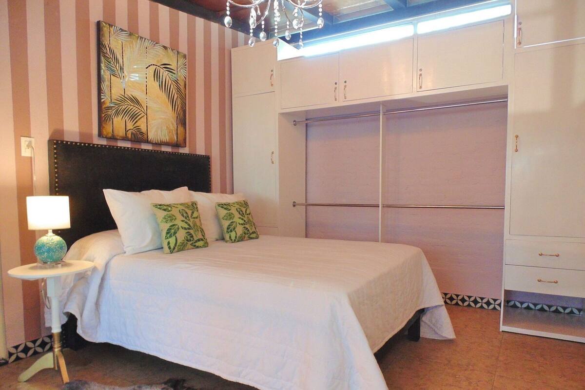 A bedroom with a bed, a nightstand, a lamp, a large artwork on the wall, and a wardrobe.