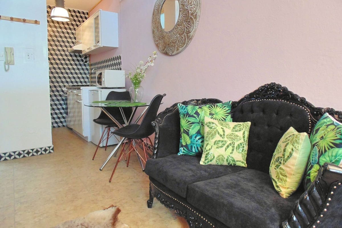 A living room with a black sofa, a dining table with chairs, a kitchen area, and a decorative mirror.
