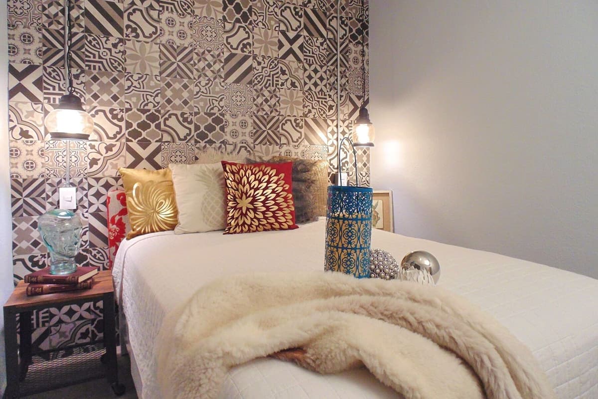 A neatly made bed with a white comforter and a fluffy white blanket, a patterned wall, and two wall-mounted lamps.