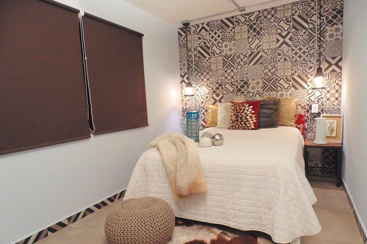 A bedroom with a patterned wallpaper, a bed with a white quilt, a brown ottoman, and a decorative rug.