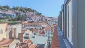A cityscape with buildings on a hillside, a clear sky, and a view from a window with blinds. - Mouraria Lisboa Hotel (Lisbon)