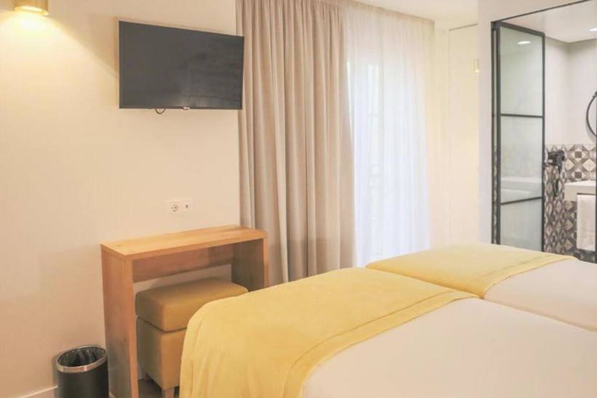 Habitación de hotel con una cama, televisor montado en la pared, una mesita de noche de madera, un cesto de basura y un baño con cabina de ducha de vidrio.