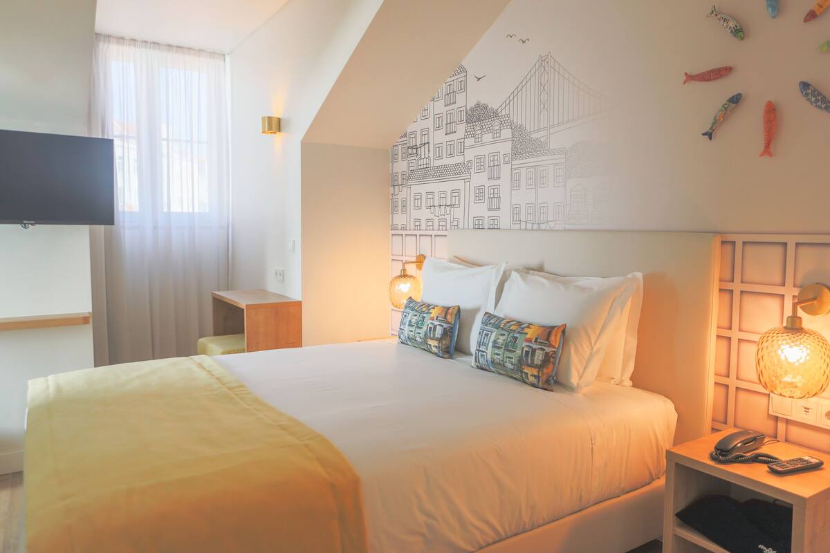 Una habitación de hotel con una cama, una mesita de noche con una lámpara, un televisor montado en la pared y un mural con edificios y peces.