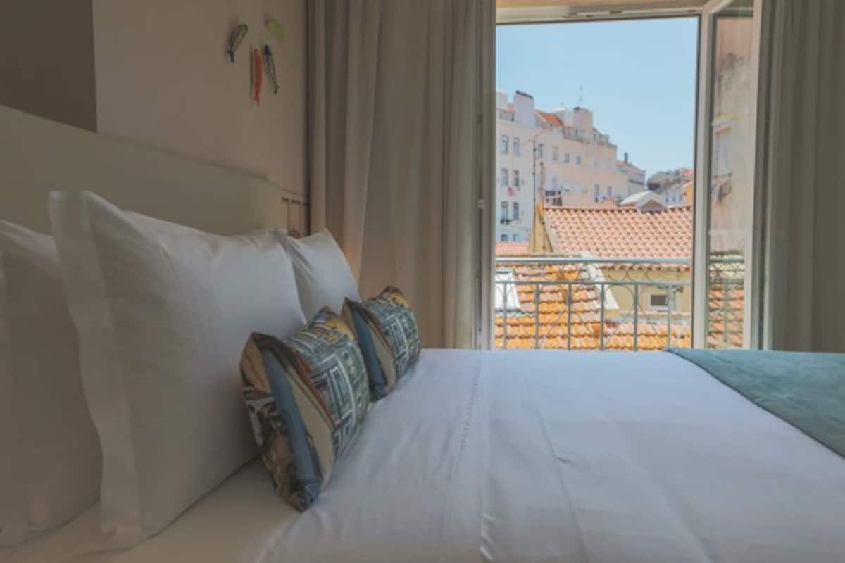 Habitación de hotel con una cama grande, vistas de edificios a través del ventanal y un balcón con barandilla.
