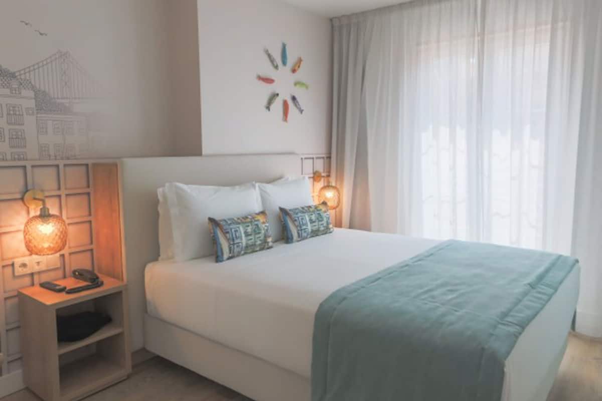 Una cama bien tendida con sábanas blancas y dos almohadones decorativos, una mesita de noche con lámpara, y una obra de arte en la pared sobre la cama.