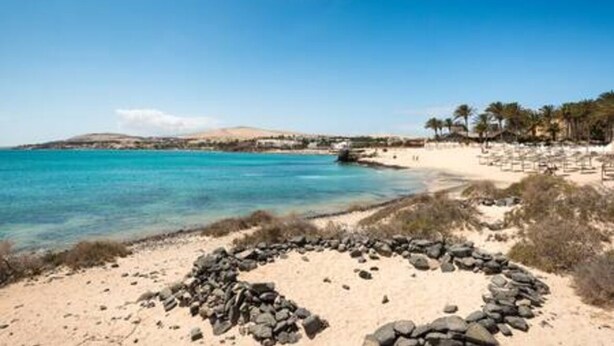 A beach with clear turquoise water, a rocky circle in the foreground, and a sandy beach with palm trees in the background. - Home2book Sea Front El Granillo, Beach & Wifi (Costa Calma)