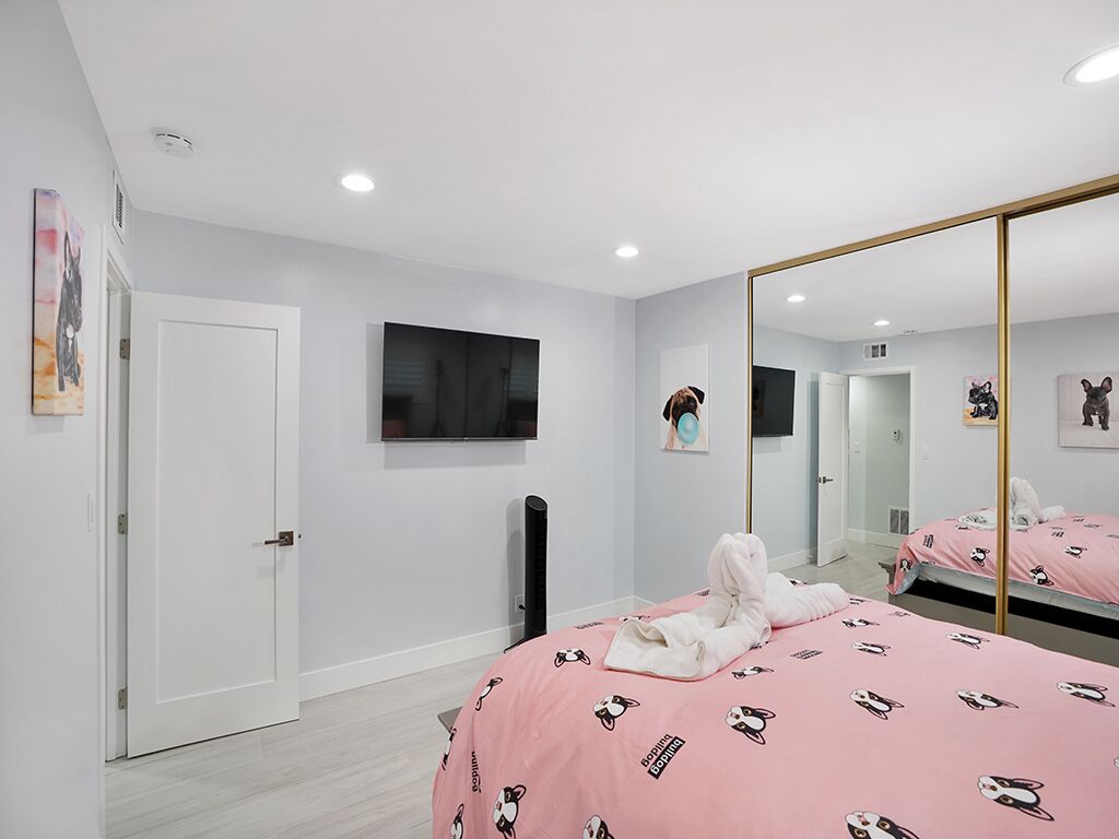 A bedroom with a bed covered in pink bedding with dog prints, a large mirror, and a flat-screen TV mounted on the wall.