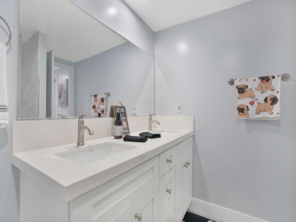 Een badkamer met een wit wastafelmeubel, twee wastafels en een grote spiegel. Er liggen handdoeken en er hangt een klein, omrande foto aan de muur.