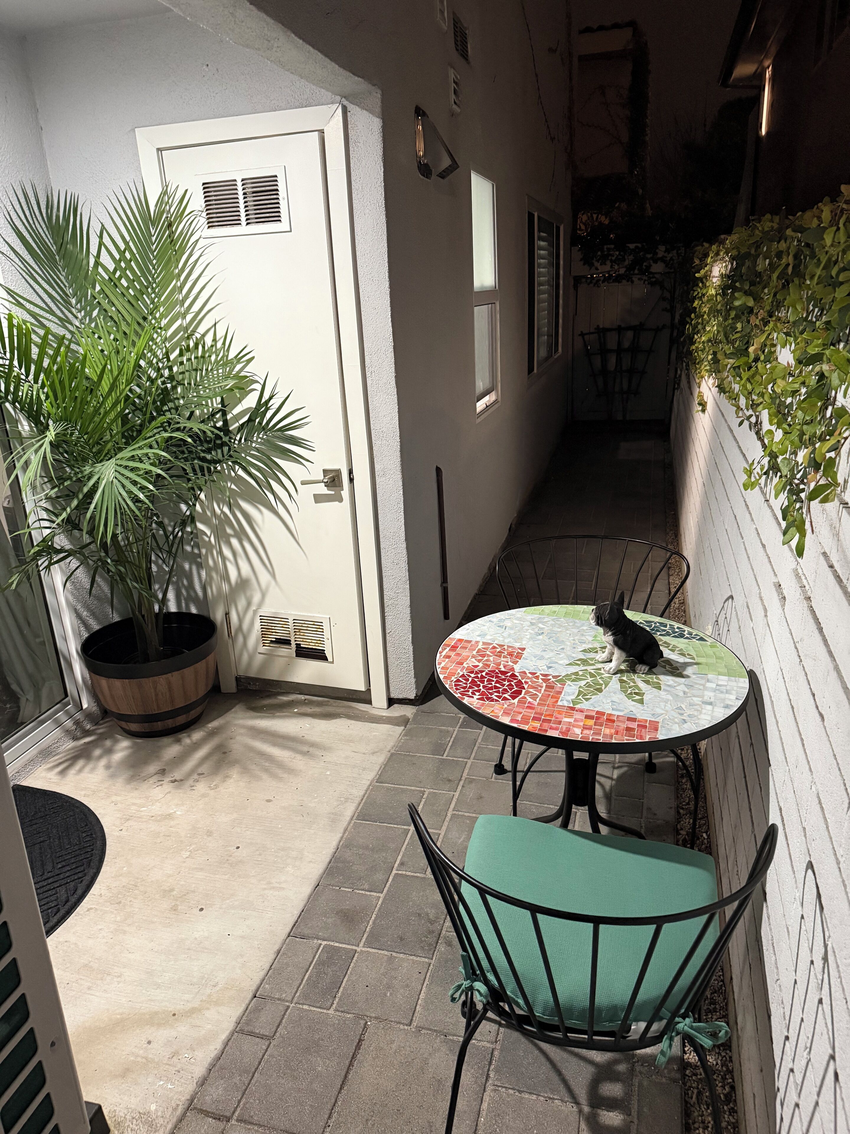 A narrow outdoor corridor with a tiled floor, a potted plant, a door, a small table with a mosaic tablecloth, and a chair.