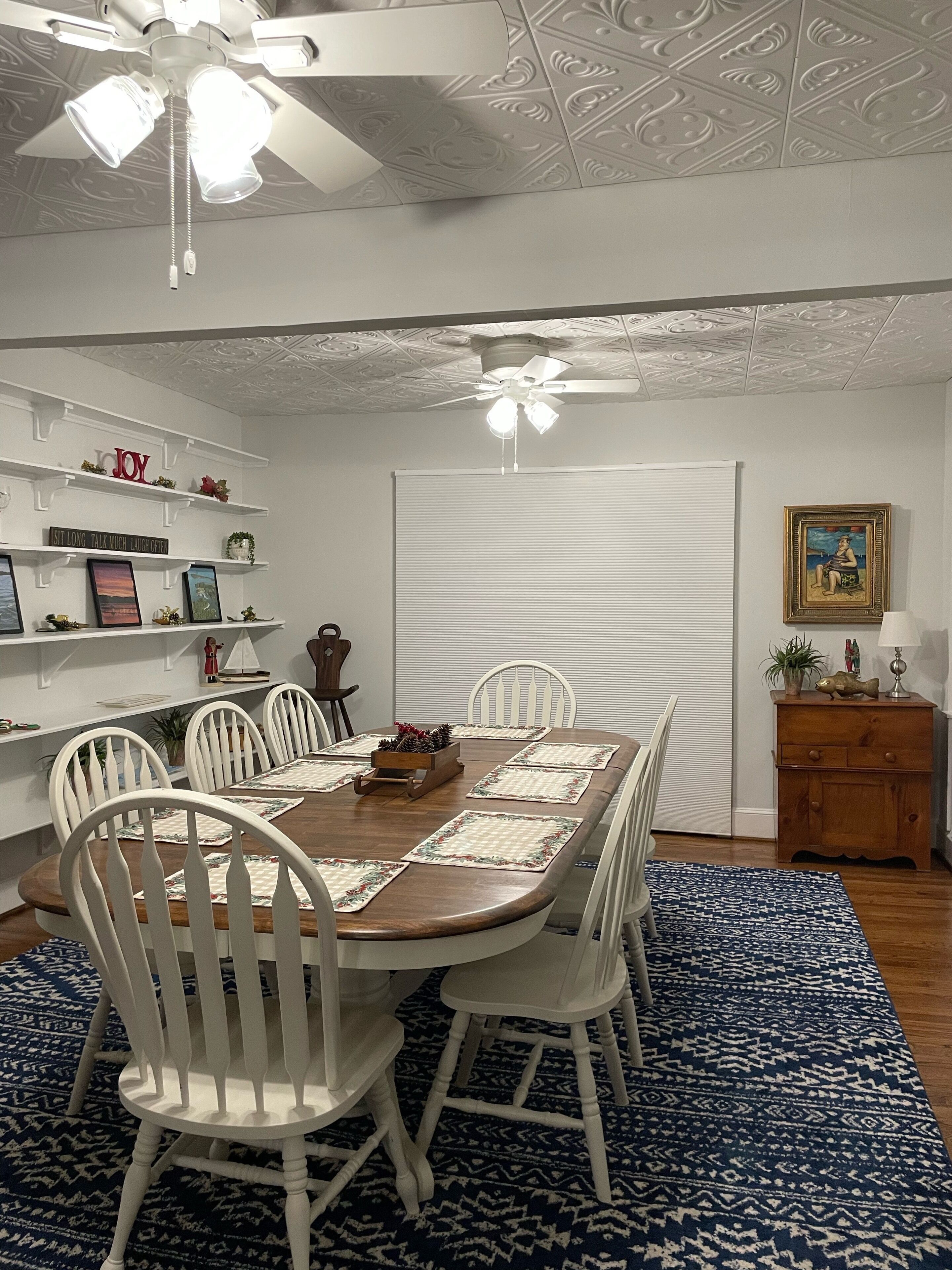 Comedor con mesa de madera, sillas blancas, alfombra azul estampada y ventilador de techo.