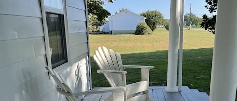 A porch with two Adirondack chairs, a wooden table, and a view of a grassy yard and a house in the background.