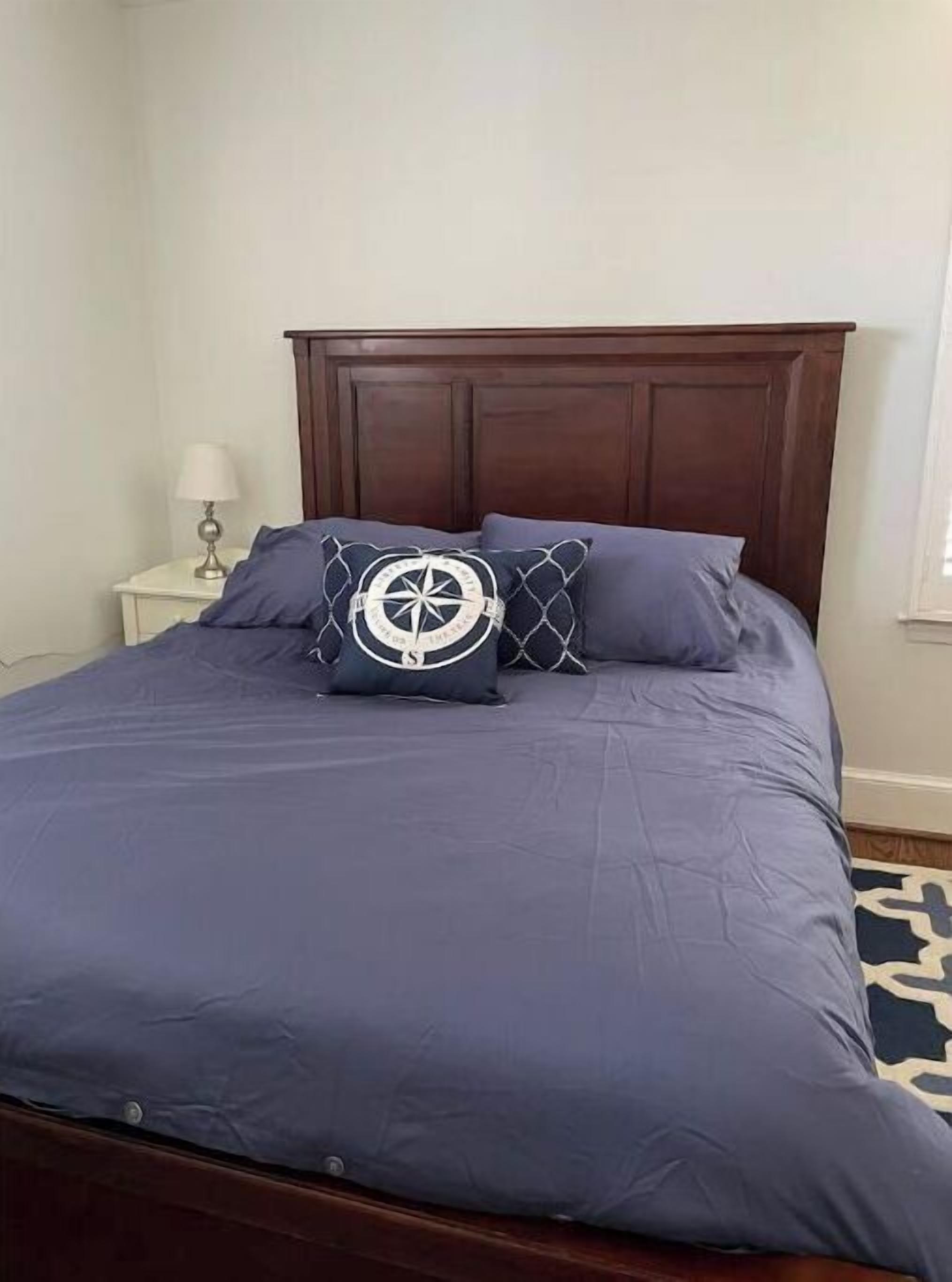Uma cama bem arrumada com um cobre-leito azul escuro e duas almofadas, uma com um desenho de bússola náutica.