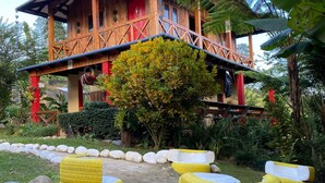 Ein Holzhaus mit roter Tür, umgeben von üppigem Grün und gelben Reifen als Sitzgelegenheit.