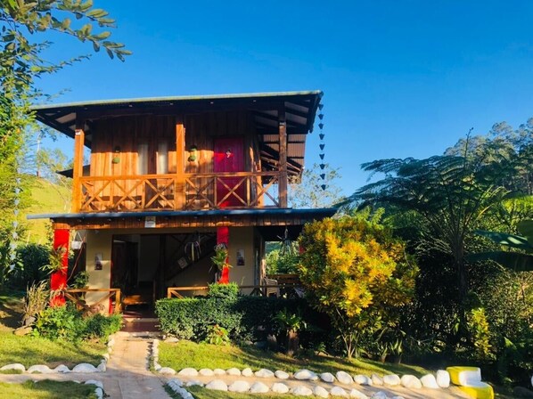 A wooden cabin with a balcony, surrounded by lush greenery and a stone pathway.
